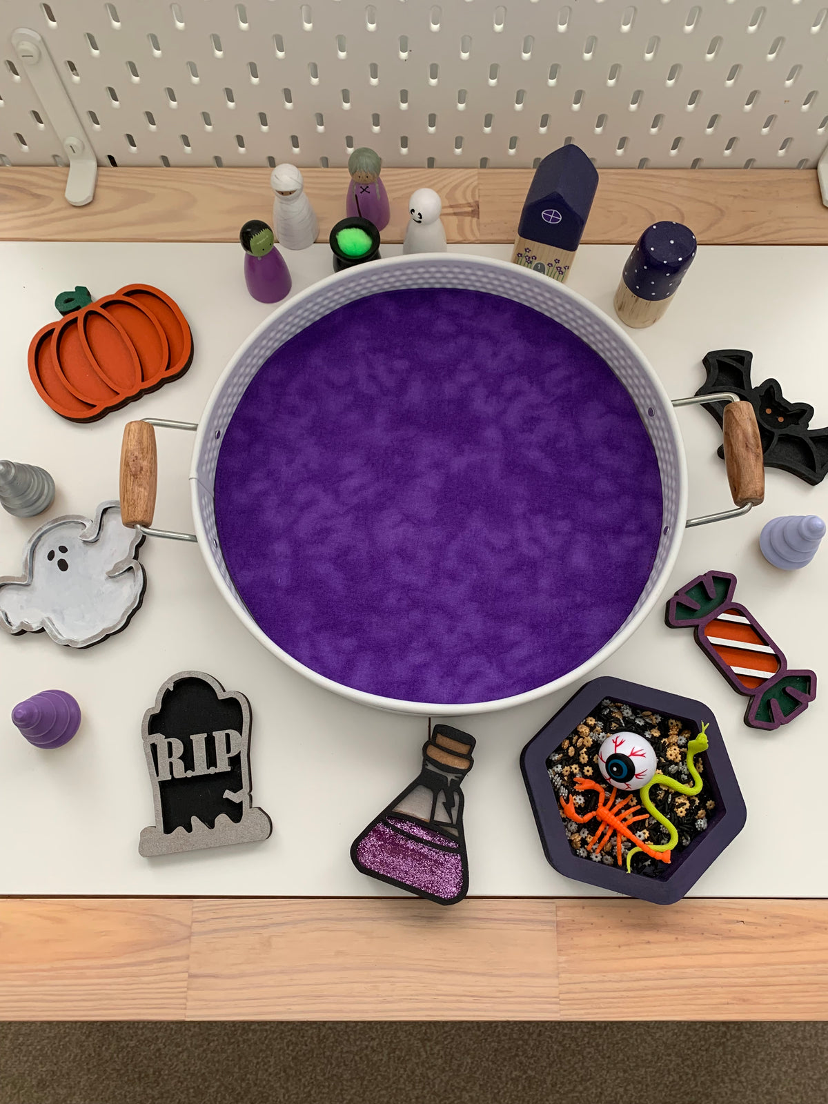 A Halloween-themed flat-lay display featuring a large white bowl filled with purple slime or paint, surrounded by various decorative craft pieces including a pumpkin, tombstone, potion bottle, candy, bats, and small figurines on a white surface.